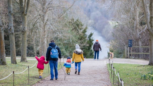 A family dressed for winter walk away from the camera down a sloping trackway flanked by trees in parkland